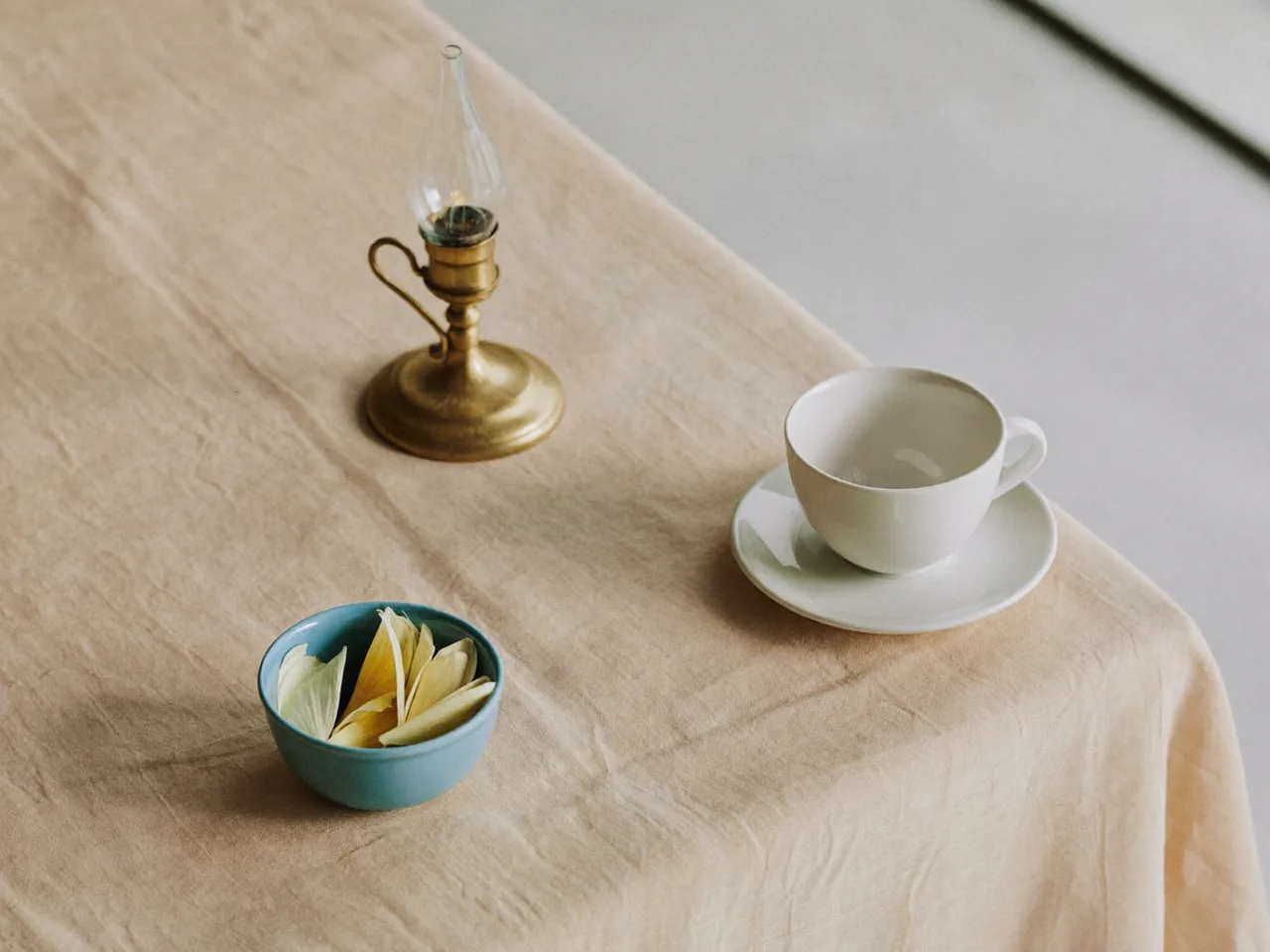 A brass lamp, a bowl of petals and a cup on a peach linen table